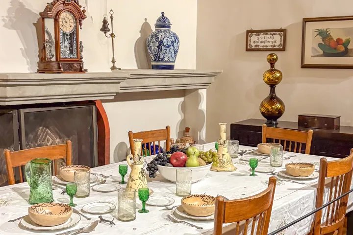 Vintage dining room with set table, wooden chairs, fruit centerpiece, decorative clock, and blue vase on mantle.