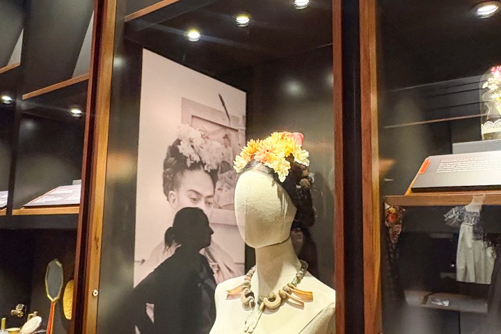 Museum display with a mannequin wearing flower crown and necklace, next to a black and white photo.