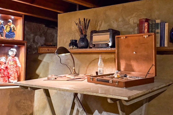 Vintage workspace with wooden desk, lamp, radio, paintbrushes, books, and display of dolls.