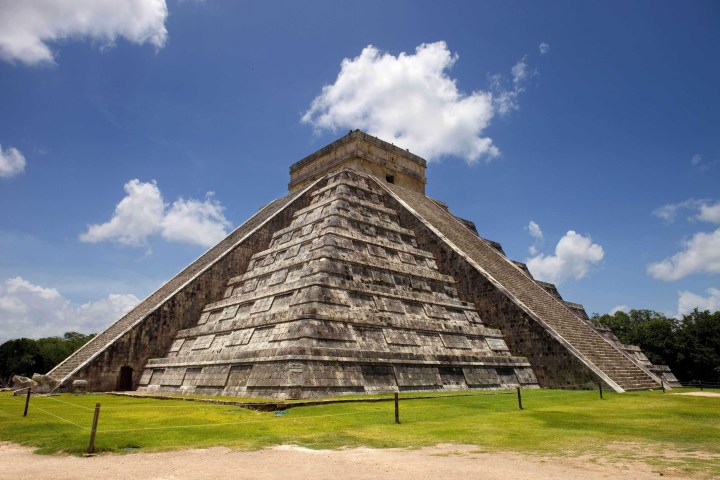 a large stone building with a grassy field with Chichen Itza in the background