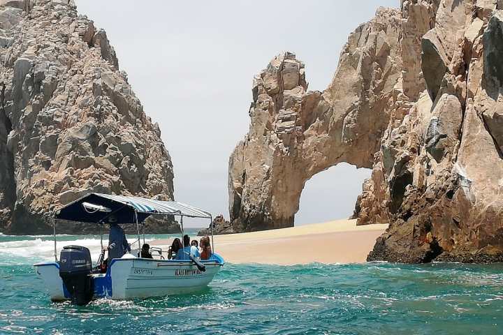 a group of people sitting on a rock near the ocean with Arch of Cabo San Lucas in the background