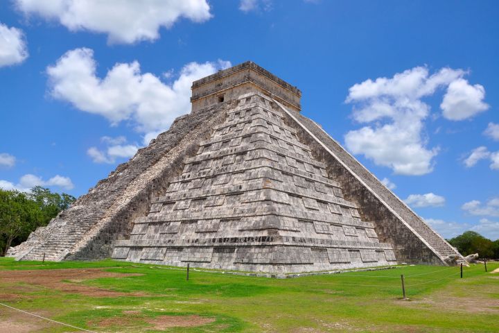 a large stone building with a grassy field with Chichen Itza in the background
