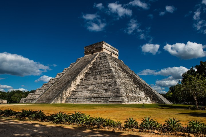 a clock tower surrounded by grass with Chichen Itza in the background