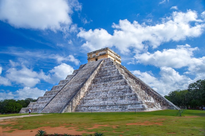a large stone building with a grassy field with Chichen Itza in the background