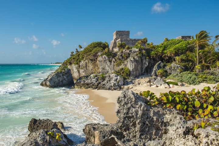 a group of people sitting on a rock near the ocean with Tulum in the background